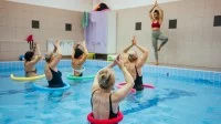 Eine Masseurin und medizinische Bademeisterin leitet vom Beckenrand aus eine Gruppe von fünf Frauen im Wasser zu gemeinsamen Yoga-Übungen an. Die Frauen sind im Schwimmbecken durchführen und haben eine Poolnudel und dem Bauch gelegt. 