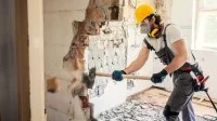Ein Bauwerksmechaniker für Abbruch und Betontrenntechnik schlägt mit einem Hammer eine Wand in einem Wohngebäude ein.