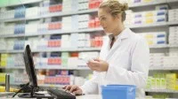 Eine pharmazeutisch-technische Assistentin im weißen Laborkittel recherchiert am Computer den Bestand eines Hautpflegeprodukts. Im Hintergrund ist ein Verkaufsregal mit verschiedenen Medikamenten.