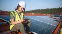 Eine technische Assistentin für regenerative Energietechnik sitzt  in Sicherheitsausrüstung mit einem Tablet in der Hand auf dem Dach neben den PV-Panelen.