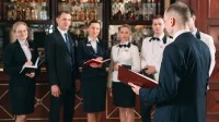 Ein Assistent für Hotelmanagement und sein Team aus sechs Mitarbeitenden stehen im Festsaal und besprechen das Menü à la carte.