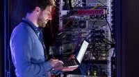 Ein Fachinformatiker arbeitet mit dem Laptop im Serverraum und überprüft die Leitungen.