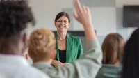 Eine lächelnde Lehrerin steht vor ihrer Klasse, ein Schüler meldet sich.
