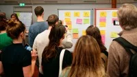 Teilnehmende auf einem Barcamp stehen vor Whiteboards auf denen verschiedene Post-it kleben.