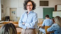 Eine Lehrerin steht mit verschränkten Armen in ihrer Klasse. Sie wirkt ruhig, gelassen und kompetent.
