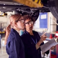 Eine Ausbilderin und Auszubildende stehen unter einem Auto auf einer hydraulischer Rampe und prüfen etwas.