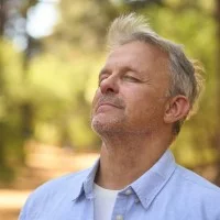 Ein Mann steht mit geschlossenen Augen auf einem Waldweg und atmet tief ein.