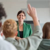 Eine lächelnde Lehrerin steht vor ihrer Klasse, ein Schüler meldet sich.