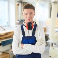 Ein Auszubildender in Arbeitsbekleidung steht mit verschränkten Armen in der Werkstatt.