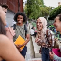 Fünf junge Menschen mit unterschiedlicher ethnischer Herkunft stehen im Kreis und lachen. Sie halten Schulsachen in den Händen.