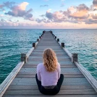 Eine Frau sitzt mit dem Rücken zum Betrachter auf einem Bootssteg und blickt zum Wasser sowie Horizont.
