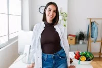 Eine Frau in einem weißen Kittel steht in ihrem Büro an einem Schreibtisch. Auf diesem steht eine Schüssel mit frischem Obst und Gemüse.