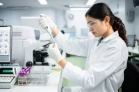 Eine medizinische Technologin in einem Laborkittel pipettiert eine Flüssigkeit in ein Reagenzglas mit einer Blutprobe. Neben ihr stehen verschiedene Analysegeräte im Labor. 