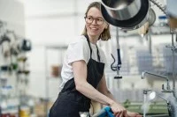 Eine Assistentin für Ernährung und Versorgung putzt die Küchenspüle in einem Heim für Menschen mit Behinderungen. Dabei trägt sie einen schwarzen Kittel und eine Brille.