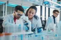 Drei biologisch-technische Assistenten arbeiten im Labor. Sie tragen weiße Kittel. Auf dem Tisch stehen ein Mikroskop und verschiedene Reagenzien. 