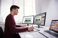 Ein Kommunikationsdesigner arbeitet mit einem Zeichenpad, zwei Bildschirmen und einem Laptop an einer neuen Werbung. 