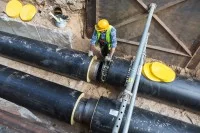 Ein Leitungsbauer steht in einer Grube, in der zwei große Rohre verlaufen. Er trägt Sicherheitskleidung und stützt sich an einem Rohr ab. 