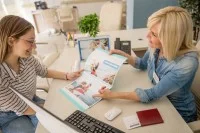 Eine Kauffrau für Tourismus und Freizeit berät im Reisebüro eine Kundin über Reiseziele am Meer.