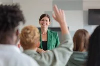 Eine lächelnde Lehrerin steht vor ihrer Klasse, ein Schüler meldet sich.