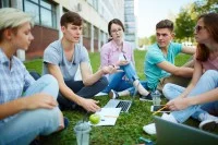 Eine Gruppe von fünf jungen Leuten sitzt bei sommerlichen Temperaturen auf einem Grünstreifen vor einem Gebäude. Die Personen haben ihre Notebooks, Schreibunterlagen sowie Getränke und Snacks dabei. 