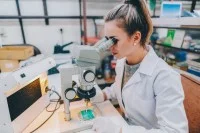 Eine junge Auszubildende sitzt an einem Mikroskop und arbeitet an einer Leiterplatte.