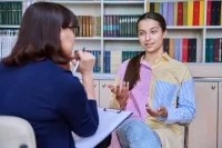 Eine Jugendliche sitzt mit einer Psychologin am Tisch, im Hintergrund ein Bücherregal.