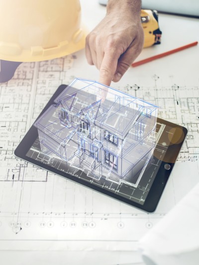 Auf einem Tisch liegt eine Bauzeichnung, auf der wiederum ein Tablet mit einem Hologramm eines Hauses sowie ein Lineal, Helm und Stift liegen. Eine Hand berührt das Hologramm.