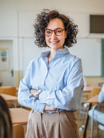 Eine Lehrerin steht mit verschränkten Armen in ihrer Klasse. Sie wirkt ruhig, gelassen und kompetent.