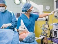 Eine Patientin in einem Krankenhaushemd wird während der Operation von einer anästhesietechnischen Assistentin betreut. Dieser überwacht die Vitalfunktionen am Monitor.