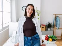 Eine Frau in einem weißen Kittel steht in ihrem Büro an einem Schreibtisch. Auf diesem steht eine Schüssel mit frischem Obst und Gemüse.