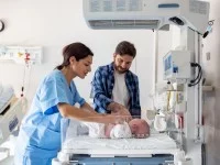 Eine Gesundheits- und Kinderkrankenpflegerin kontrolliert den Gesundheitszustand eines Neugeborenen. Daneben steht der Vater des Babys, der mit seiner Hand das Baby streichelt.