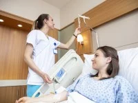 Eine Gesundheits- und Krankenpflegerin überprüft die Einstellung des Tropfes einer Patientin. Diese liegt im Krankenzimmer in ihrem Krankenbett.