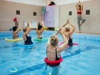 Eine Masseurin und medizinische Bademeisterin leitet vom Beckenrand aus eine Gruppe von fünf Frauen im Wasser zu gemeinsamen Yoga-Übungen an. Die Frauen sind im Schwimmbecken durchführen und haben eine Poolnudel und dem Bauch gelegt. 