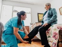 Eine Pflegefachfrau hilft einer älteren Frau beim Anziehen ihres Schuhs. Die ältere Frau sitzt dabei in ihrem Schlafzimmer auf ihrem Bett. 