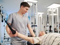 Ein Physiotherapeut unterstützt einen älteren Herrn in einem Fitnessraum dabei, seine Kraft in den Armen zu trainieren. 