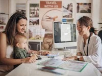 Eine Touristikassistentin berät eine junge Mutter mit Kleinkind auf dem Schoß am Schreibtisch über die Urlaubsplanung.