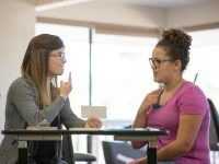 Eine Atem-, Sprech- und Stimmlehrerin leitet eine Patientin zu einer besseren Stimmbildung an. Beide Frauen sitzen dabei am Tisch. 