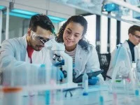 Drei biologisch-technische Assistenten arbeiten im Labor. Sie tragen weiße Kittel. Auf dem Tisch stehen ein Mikroskop und verschiedene Reagenzien. 