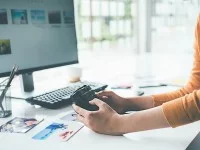 Hände halten eine Kamera vor einem Computerbildschirm. Auf dem Tisch befinden sich Fotos, eine Tastatur und ein Stiftehalter.