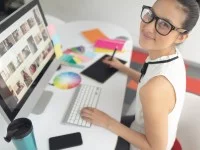 Eine glückliche gestaltungstechnische Assistentin arbeitet an ihrem Computer. Auf dem Bildschirm werden verschiedene Fotos angezeigt, zudem liegen Farbtafeln auf ihrem Schreibtisch. 