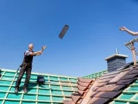 Ein Dachdecker wirft seinem Kollegen auf dem Dach einen Ziegel zu.