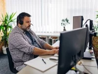 Ein übergewichtiger Mann mit einem Headset auf dem Kopf sitzt im Büro an seinem Schreibtisch und tippt etwas in die Computertastatur. 