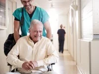 Ein glücklicher Gesundheits- und Krankenpflegehelfer fährt einen älteren Mann im Rollstuhl über den Flur und trägt dabei die Tasche des Mannes.