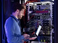 Ein Fachinformatiker arbeitet mit dem Laptop im Serverraum und überprüft die Leitungen.